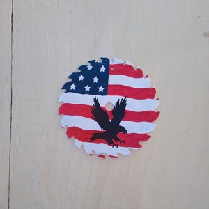 May include: A circular saw blade painted with the American flag design. The blade features red and white stripes, a blue field with white stars, and a black eagle silhouette. The saw blade is displayed on a light wood surface.