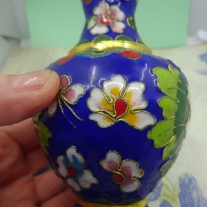 May include: A small blue and gold cloisonné vase with a floral design. The vase is decorated with white, red, yellow, and green flowers.