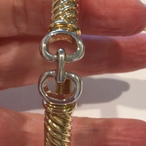 May include: Gold-toned bracelet with a twisted rope design. The bracelet features a silver-colored clasp with a unique interlocking design. The jewelry is a classic accessory.