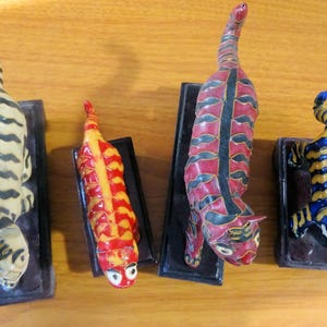 May include: Four ceramic figurines of cats, each with a different color pattern. The cats are standing on wooden bases and have a whimsical, stylized design.