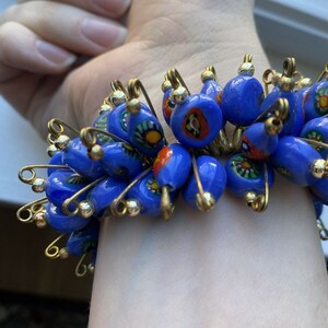 May include: A blue beaded bracelet with gold-colored safety pins. The beads are a vibrant blue with colorful floral designs. The bracelet is worn on a wrist.