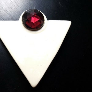 May include: A white triangular brooch with a faceted, dark red gemstone at the top. The brooch has a simple, geometric design and is set against a dark background. This vintage jewelry piece is a unique accessory.