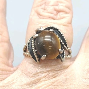 May include: A silver ring with a brown tiger's eye stone set in a twisted silver band. The band is decorated with black metal leaves.