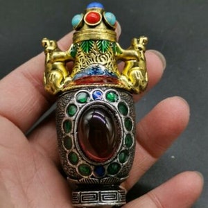 May include: An ornate, antique bottle with a silver base and gold accents. The bottle features a large, oval, red gemstone and is adorned with green, blue, and red jewels. Two gold lion figures are on the top.