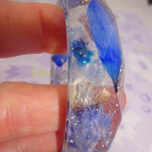 Op de afbeelding: Een transparante hars armband met ingebedde blauwe en witte geperste bloemen en glitter.
