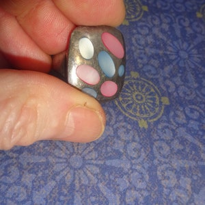 May include: A silver-toned ring with a rectangular face. The face features an arrangement of oval-shaped inlays in pink, light blue, and white. The ring is held by a hand against a blue patterned background.