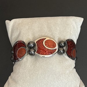 May include: A silver bracelet with red and orange enamel accents. The bracelet features a geometric design with rounded edges.