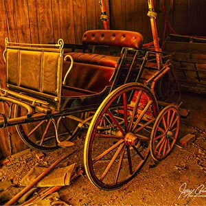 May include: A vintage horse-drawn carriage with a brown leather seat and wooden wheels. The carriage is in a barn with a wooden wall in the background. The carriage is in a state of disrepair, with some of the wood showing signs of wear and tear. The carriage is a dark brown color and the wheels are a lighter brown color. The carriage is a classic example of a horse-drawn carriage from the early 20th century.