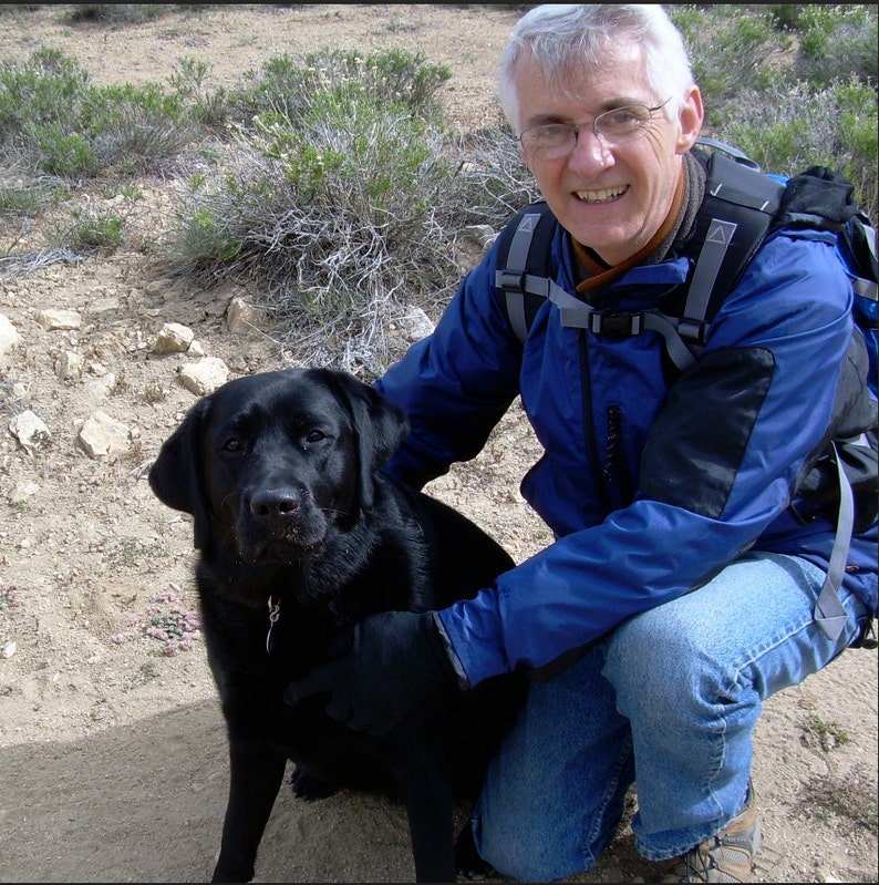 May include: A man wearing a blue jacket and a backpack is kneeling down next to a black Labrador Retriever.