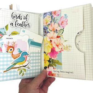 May include: An open scrapbook with a blue and white checkered pocket on the left side. The pocket contains a sticker of a blue bird on a branch, a sticker of a deer, and a sticker that says "FRIENDS". The right side of the scrapbook has a floral design with the text "Spring feels magical."