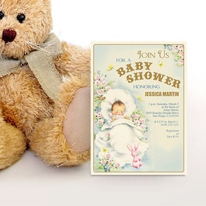 Puede incluir: Un conejito de peluche marr&oacute;n con una bufanda gris se sienta junto a una invitaci&oacute;n a un baby shower. La invitaci&oacute;n es para Jessica Martin y presenta a un beb&eacute; en una manta blanca con un conejito rosa. La invitaci&oacute;n incluye la fecha, la hora y la ubicaci&oacute;n de la ducha.