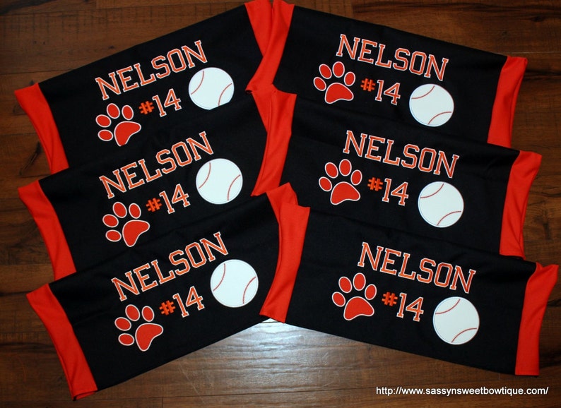 Puede incluir: Seis brazaletes de tela negra y naranja con el nombre "NELSON" y el n&uacute;mero "14" impresos en blanco. Cada brazalete tambi&eacute;n tiene una huella de pata roja y una pelota de b&eacute;isbol blanca.