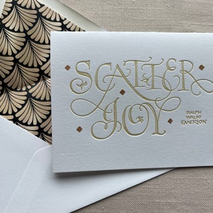 May include: A white card with gold lettering that reads "Scatter Joy" and a quote from Ralph Waldo Emerson. The card is in a white envelope with a black and gold patterned lining.