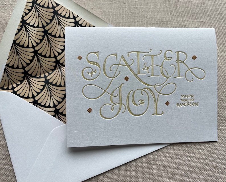 May include: A white card with gold lettering that reads "Scatter Joy" and a quote from Ralph Waldo Emerson. The card is in a white envelope with a black and gold patterned lining.