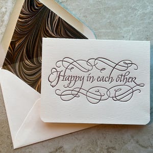 May include: A white card with a decorative script font that reads "Happy in each other". The card is sitting on top of a white envelope with a brown and black marbled pattern.