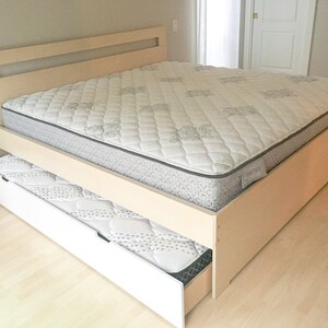 May include: A wooden bed frame with a white mattress and a pull-out drawer with a mattress. The bed frame has a light wood finish and a headboard with a rectangular cutout.