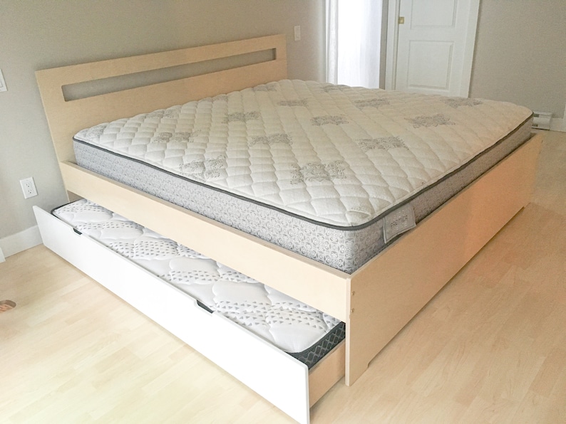 May include: A wooden bed frame with a white mattress and a pull-out drawer with a mattress. The bed frame has a light wood finish and a headboard with a rectangular cutout.
