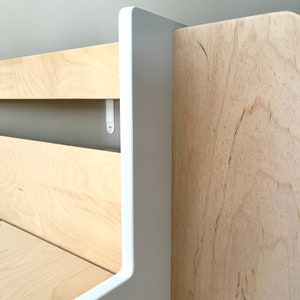 May include: Close-up of a white and natural wood shelf unit with a rounded edge. The shelf is made of wood with a natural grain pattern. The shelf is attached to a wall with white brackets.