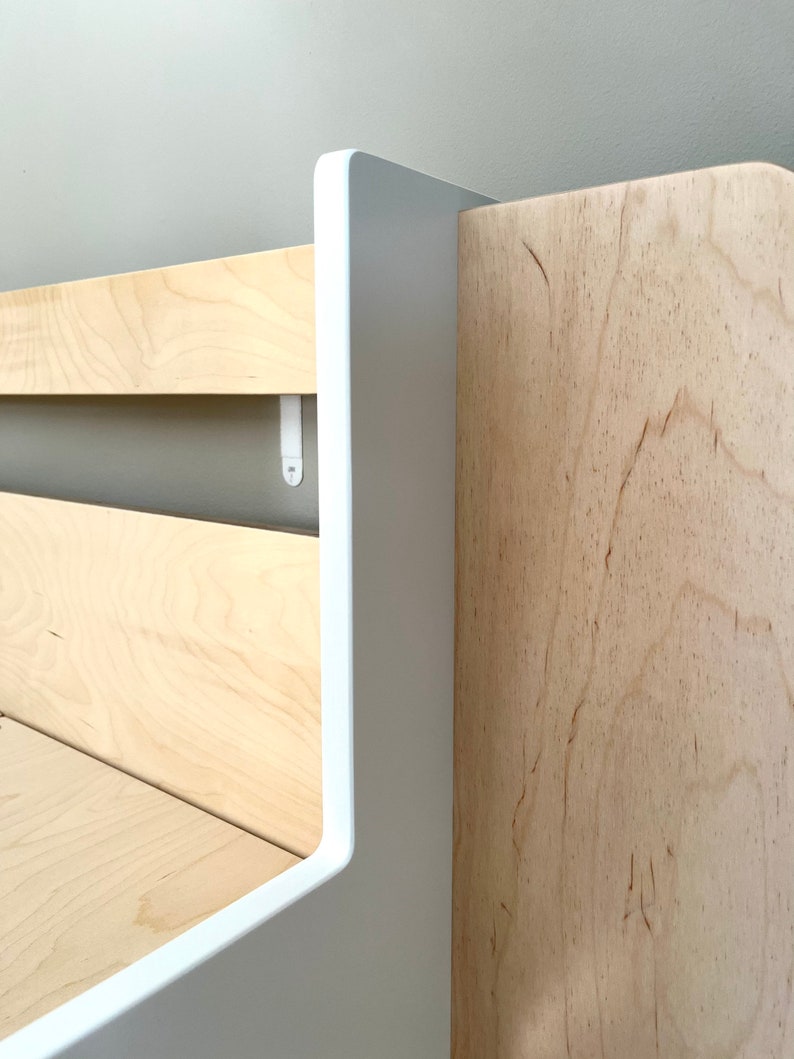 May include: Close-up of a white and natural wood shelf unit with a rounded edge. The shelf is made of wood with a natural grain pattern. The shelf is attached to a wall with white brackets.