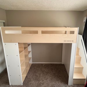 May include: A white and wood loft bed with a built-in bookshelf and stairs. The bed frame is made of wood and has a white finish. The bookshelf has multiple shelves and is located on the left side of the bed. The stairs are made of wood and have a white finish. The bed frame has a cutout in the headboard that says "DEDRICK".