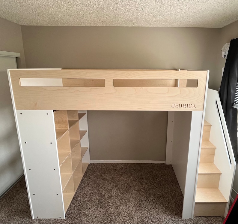 May include: A white and wood loft bed with a built-in bookshelf and stairs. The bed frame is made of wood and has a white finish. The bookshelf has multiple shelves and is located on the left side of the bed. The stairs are made of wood and have a white finish. The bed frame has a cutout in the headboard that says "DEDRICK".