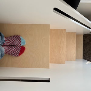 May include: A set of wooden steps with a light wood finish. The steps are attached to a white wall. A person's feet are visible at the bottom of the steps, wearing red and blue patterned socks.