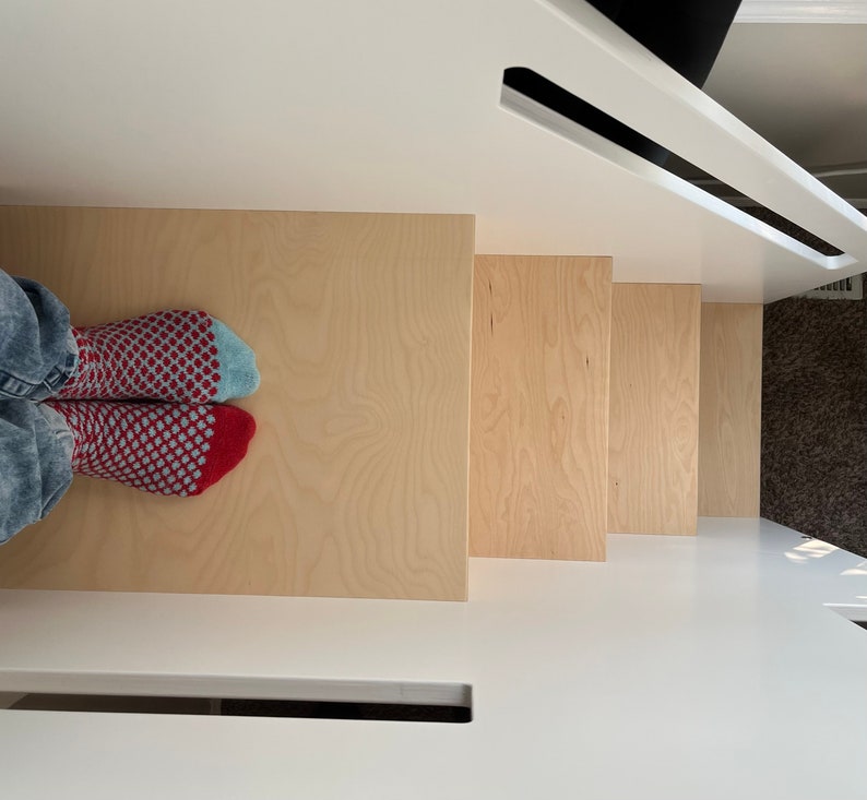 May include: A set of wooden steps with a light wood finish. The steps are attached to a white wall. A person's feet are visible at the bottom of the steps, wearing red and blue patterned socks.