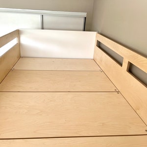 May include: A close-up of a wooden bed frame with a light wood finish. The bed frame has a simple design with a few slats for support. The bed frame is made of plywood and has a natural wood grain.