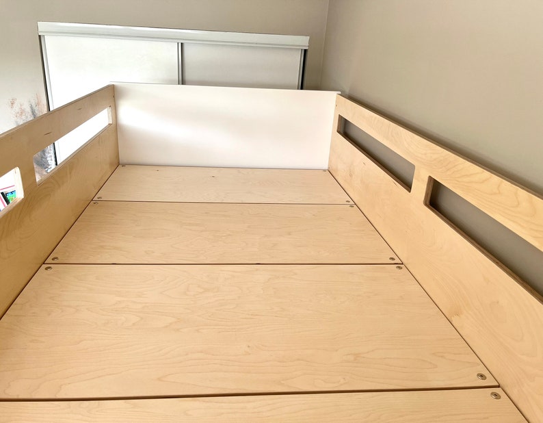May include: A close-up of a wooden bed frame with a light wood finish. The bed frame has a simple design with a few slats for support. The bed frame is made of plywood and has a natural wood grain.
