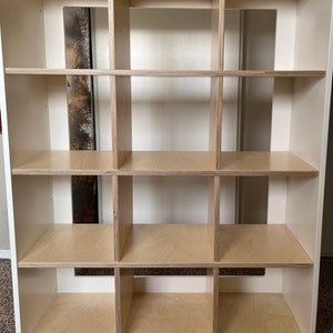May include: White cube shelf unit with 12 compartments. The shelves are made of light wood and the frame is white.