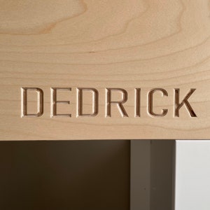 May include: A close-up of a light wood surface with the name "Dedrick" carved into it.