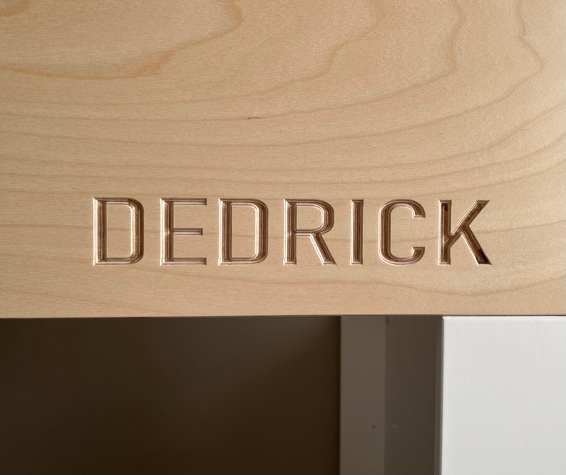 May include: A close-up of a light wood surface with the name "Dedrick" carved into it.