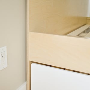 May include: A close-up of a wooden bed frame with a white drawer. The bed frame is made of plywood and has a simple, modern design.