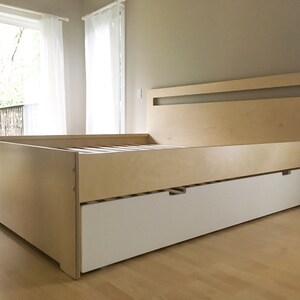 May include: A light wood platform bed frame with a white drawer underneath. The headboard has a rectangular cutout in the top.