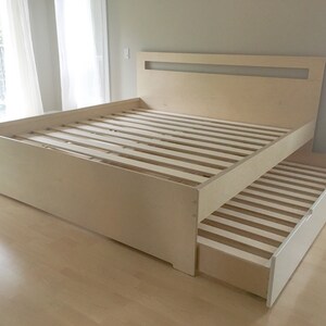 May include: A light wood platform bed frame with a built-in headboard and a pull-out trundle bed. The bed frame has a simple, modern design.