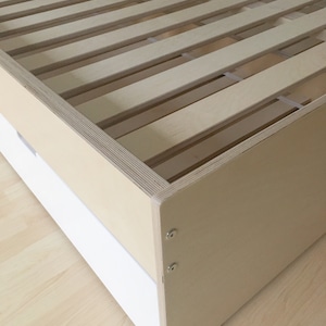 May include: A wooden bed frame with a slatted base and a white drawer. The frame is made of plywood and has a light brown finish. The drawer is attached to the front of the frame and has a white finish.