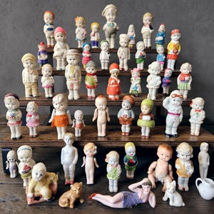 May include: A collection of antique bisque dolls of various sizes and styles, arranged on wooden shelves. The dolls are dressed in colorful outfits, with painted faces and detailed features. Some dolls are nude, while others are holding accessories.