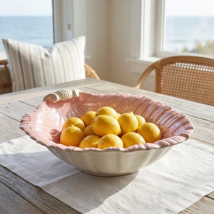 May include: A large, scalloped ceramic bowl shaped like a seashell, featuring a glossy pastel pink interior and a cream-colored exterior. The bowl is filled with a cluster of bright yellow lemons, showcasing a textured, ribbed surface design.