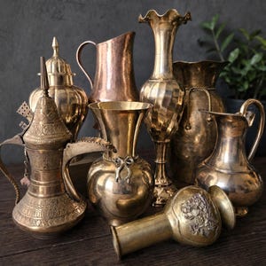 May include: A collection of various brass and copper vessels, including ornate pitchers, tall vases with fluted or smooth bodies, a lidded container, and a small vase with floral relief detail, all featuring metallic finishes and distinct shapes.