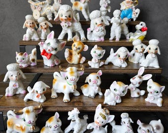 May include: A collection of vintage ceramic figurines, including lambs, poodles, cats, a bear, a cow, a pig, and a rabbit. The figurines are primarily white with pastel accents and are arranged on wooden shelves.