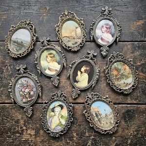 May include: A collection of eight oval-shaped picture frames with ornate, metallic bronze-toned filigree borders. Each frame contains a distinct miniature print, including portraits of people, a floral arrangement, and various landscape scenes.