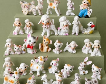 May include: A collection of small ceramic animal figurines including poodles, cats, cows, and a child in a yellow hat. The figures feature painted details like pink flowers, red bows, and gold accents, arranged on a tiered green display stand.