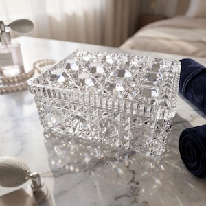 May include: A rectangular clear glass jewelry box featuring a faceted, diamond-like geometric pattern across the lid and sides. The box has a thick, textured rim and a transparent, reflective surface that catches light to create sharp, angular highlights.