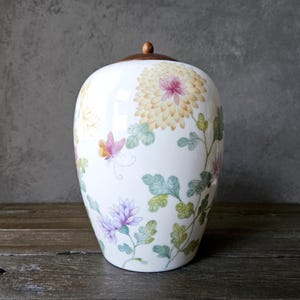 May include: A white ceramic jar with a wooden lid, adorned with floral and butterfly designs in yellow, pink, and purple. The jar has a rounded shape and is decorated with green leaves and stems.