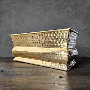 May include: A rectangular, hammered brass planter with a gold finish. The planter has a textured surface and a curved base. The planter is approximately 12 inches long, 6 inches wide, and 5 inches tall.