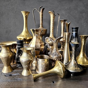 May include: A collection of antique brass vases and pitchers in various shapes and sizes. Some have handles, while others are ornate with etched designs. The vases are arranged on a wooden surface, reflecting a vintage aesthetic.