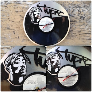 May include: A round wall clock featuring a black vinyl record with a silver-colored metal frame. The clock has a black and white image of Tupac Shakur and the word "tupac" in black. Red clock hands indicate the time.