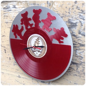 May include: A red and silver vinyl record clock featuring a Grateful Dead logo and dancing bears. The clock has a silver background with red dancing bears and a red vinyl record with a Grateful Dead logo in the center.