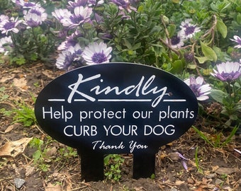 Op de afbeelding: Een zwart metalen bord met witte tekst die "Kindly Help protect our plants CURB YOUR DOG Thank You" luidt. Het bord heeft de vorm van een ovaal en is in de grond gestoken voor een bed met paarse bloemen.