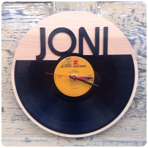 May include: A round wall clock made from a vinyl record, featuring the name "JONI" in black letters above the record. The clock face includes a record label and red and black clock hands. The clock is mounted on a light wood background.